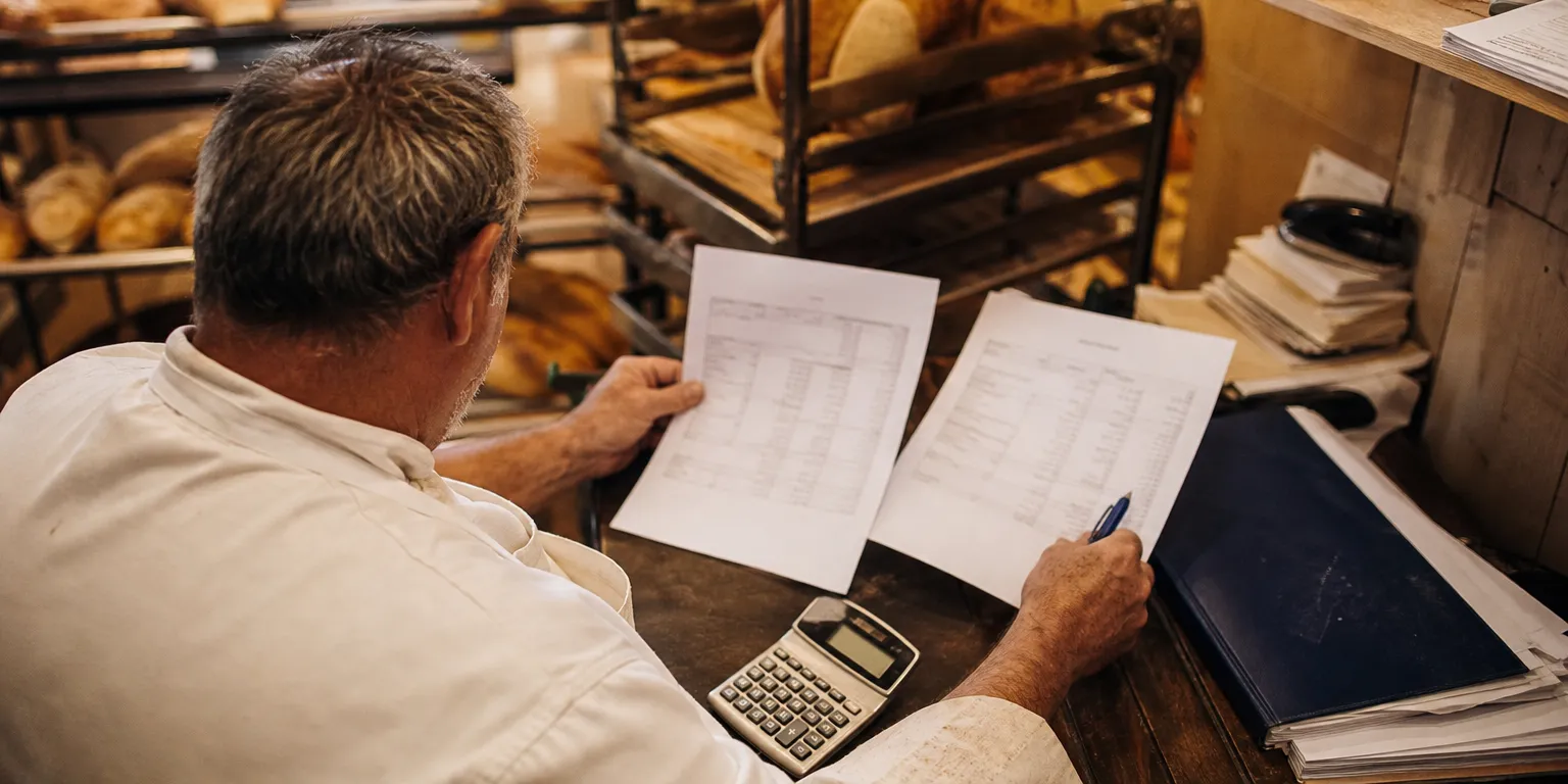 Artisan boulanger comparant ses relevés annuels de frais bancaires dans son arrière-boutique